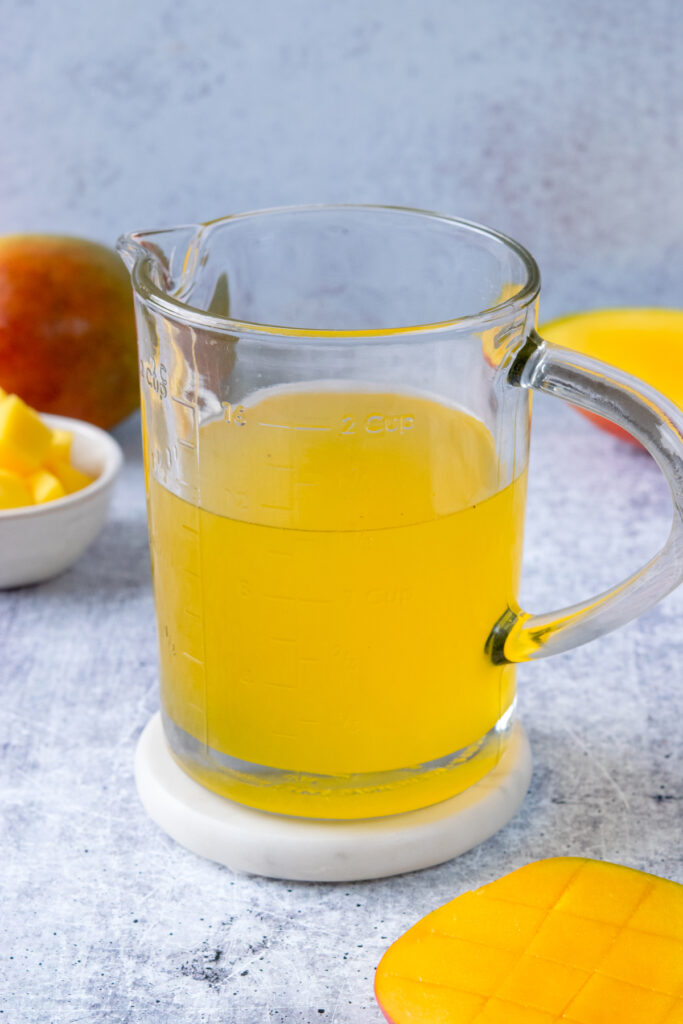 Bright yellow homemade mango syrup in a small glass pitcher with a scored mango half and a whole mango in the background.