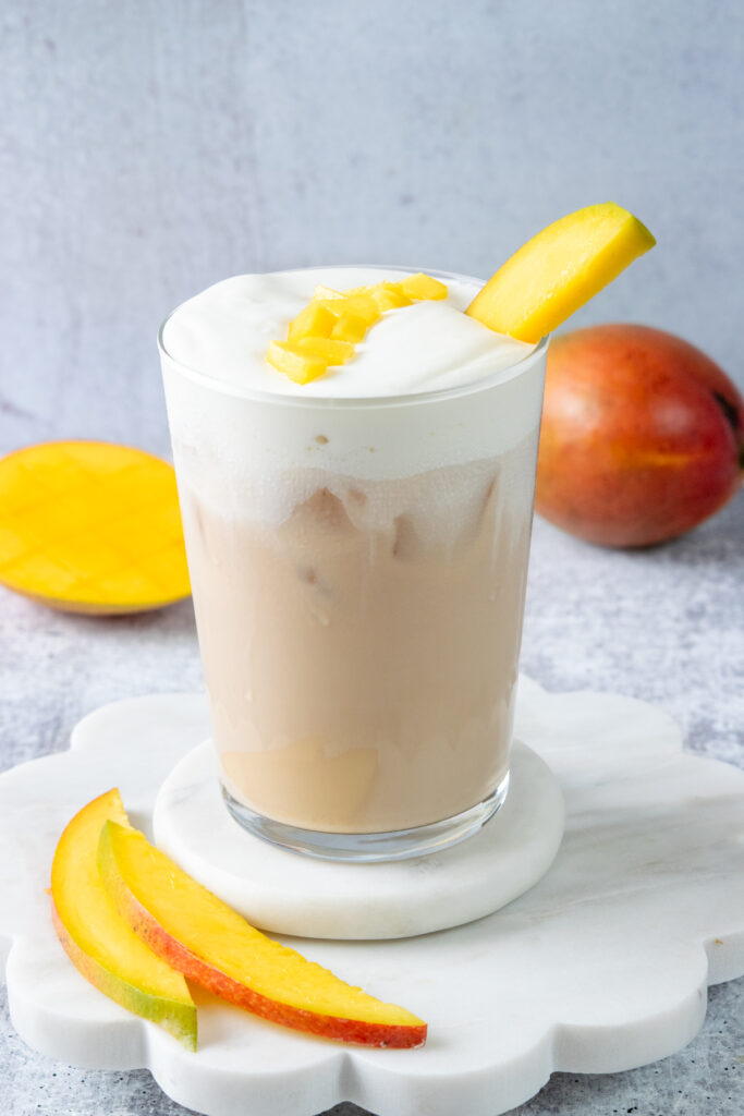 A creamy iced mango cream chai in a glass, topped with mango cold foam and garnished with fresh mango chunks and a slice of mango.