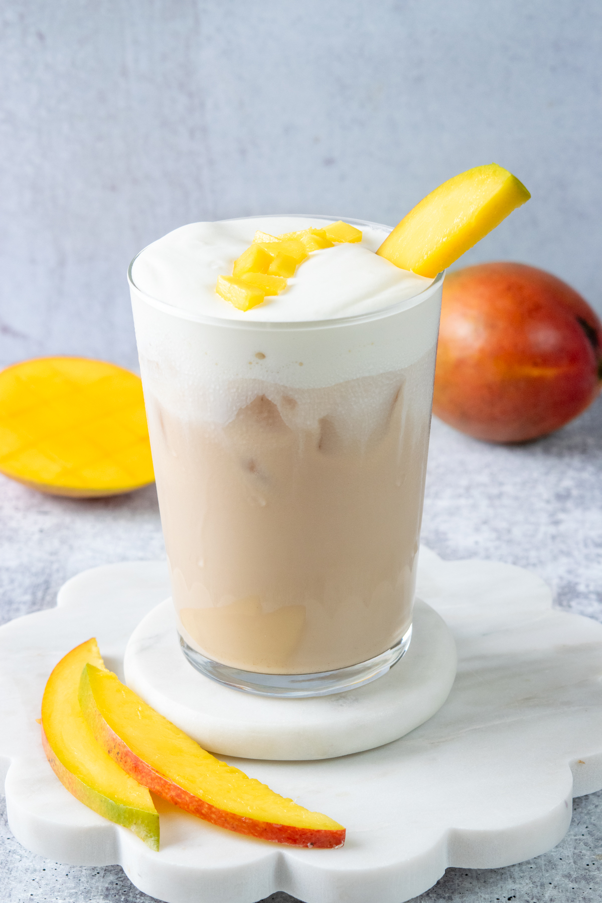A creamy iced mango cream chai in a glass, topped with mango cold foam and garnished with fresh mango chunks and a slice of mango.