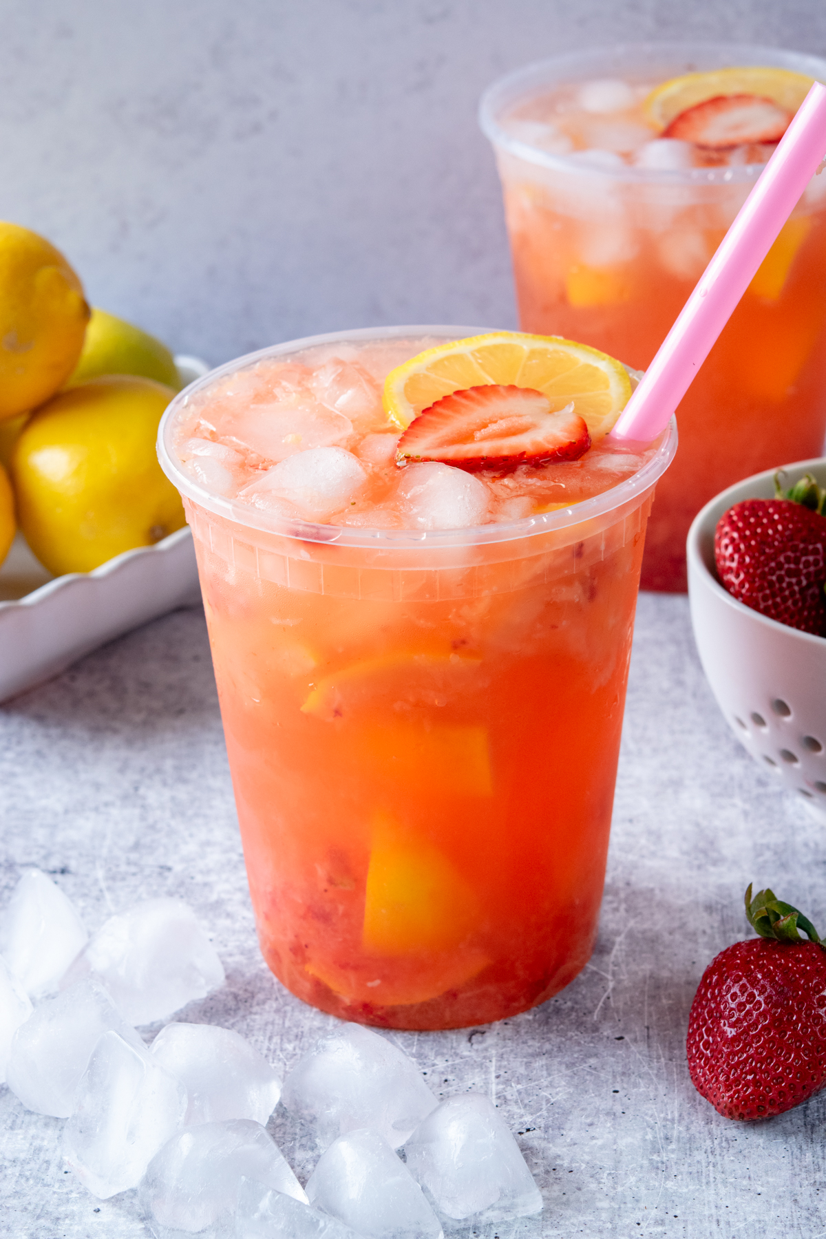A refreshing state fair strawberry lemonade in a 32 ounce deli container filled with ice, with another shake-up lemonade, fresh lemons, and strawberries in a berry bowl in the background.