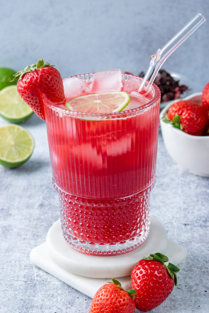 A glass of ice-cold strawberry hibiscus iced tea garnished with fresh lime and a strawberry, surrounded by strawberries, dried hibiscus flowers, and halved limes for a refreshing summer drink scene.