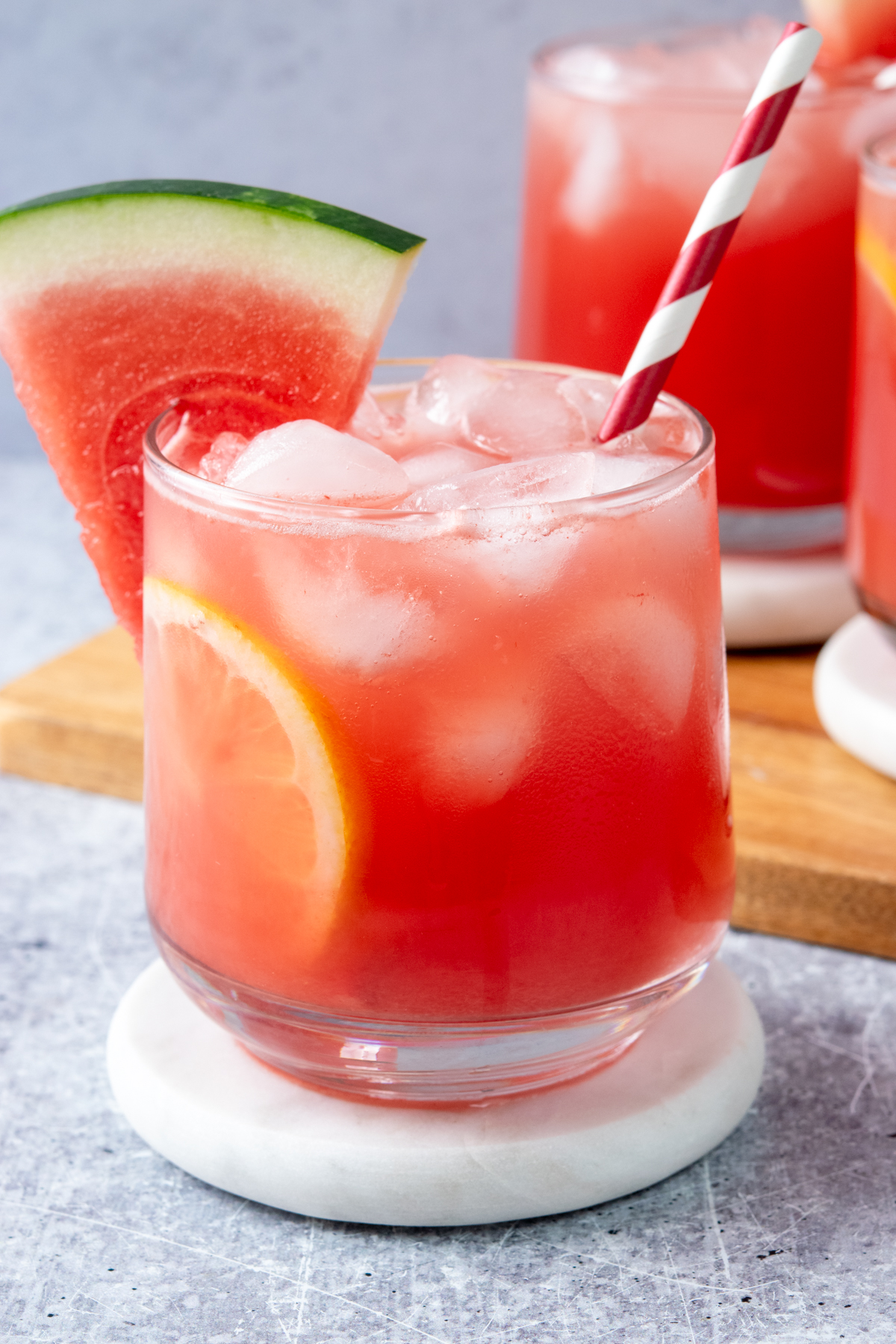 Three glasses of ice-cold watermelon lemonade filled with ice and garnished with lemon wheels, watermelon wedges, and straws for a refreshing summer drink.