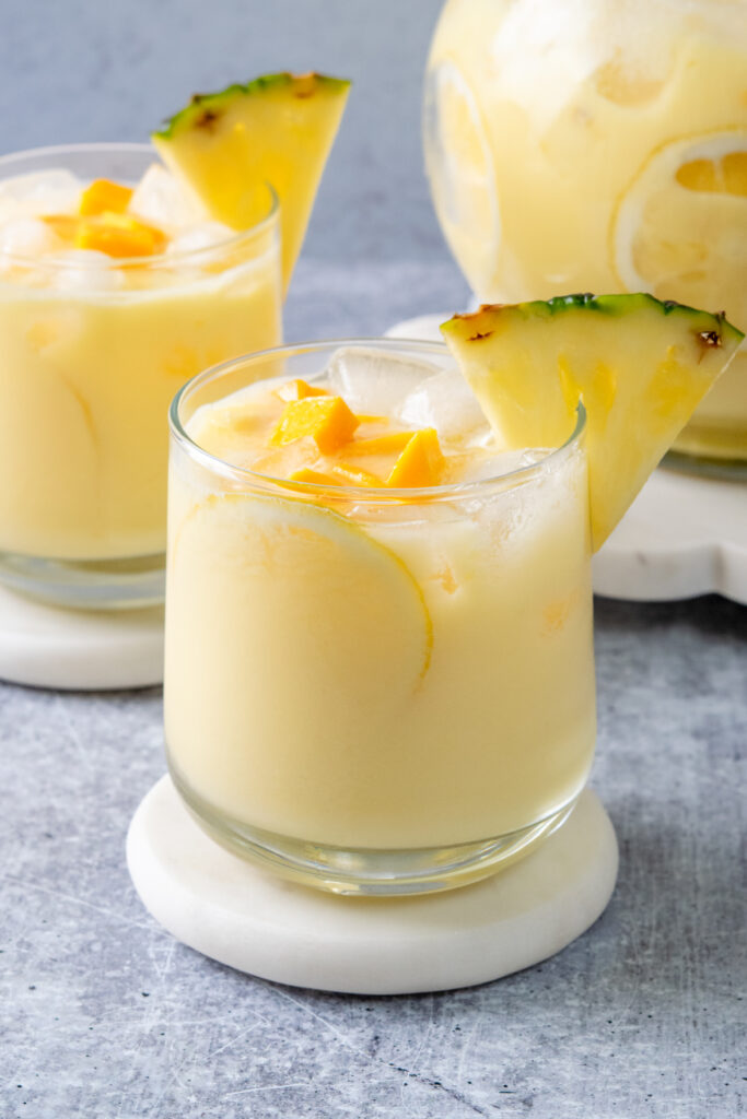 Two glasses of creamy pineapple mango lemonade served with a pitcher, garnished with lemon slices, fresh pineapple wedges, and juicy mango chunks.