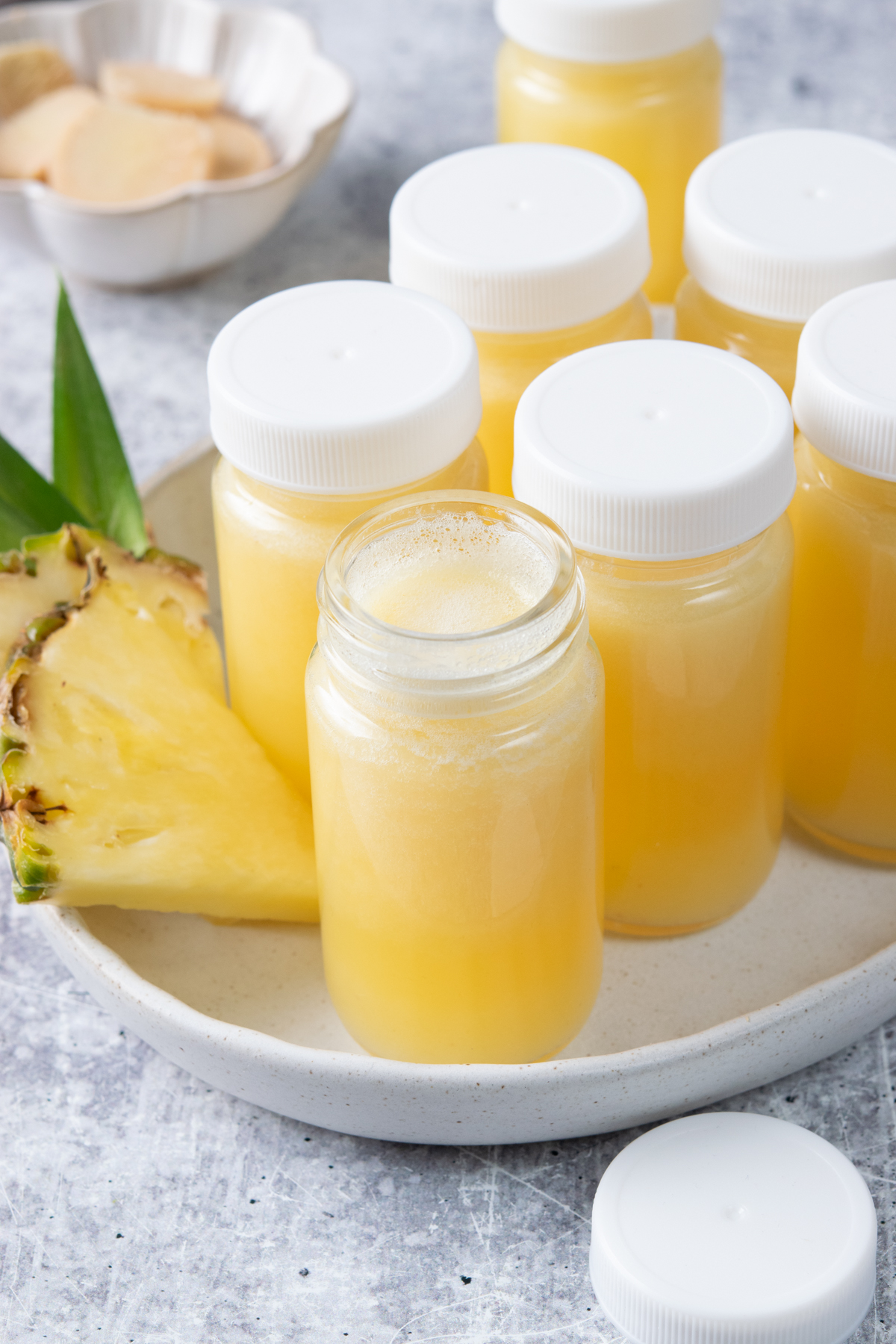 Tray of homemade pineapple ginger shots in small glass shot bottles, with one bottle open in front. The bright yellow shots are surrounded by fresh pineapple slices and ginger root.