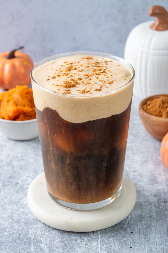 A copycat Starbucks pumpkin cream cold brew topped with thick pumpkin cold foam and pumpkin pie spice. The iced coffee is served in a clear glass with a small pumpkin and bowl of pumpkin puree behind it.