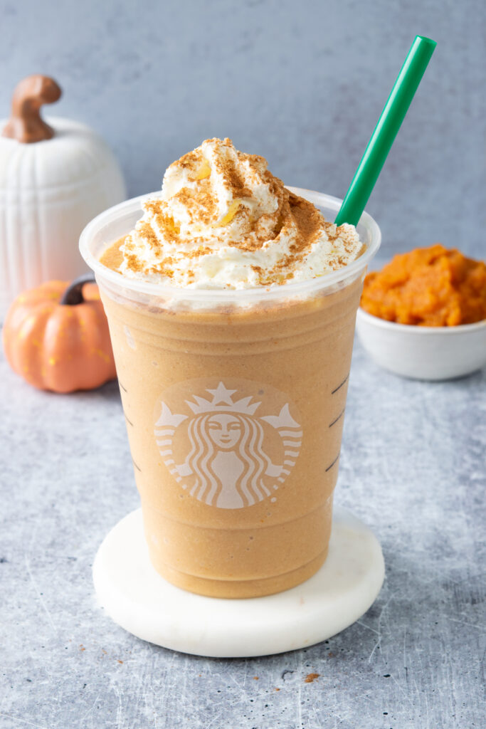 Homemade Starbucks copycat pumpkin spice frappuccino topped with whipped cream and pumpkin pie spice, served beside a small bowl of pumpkin puree and a decorative pumpkin.