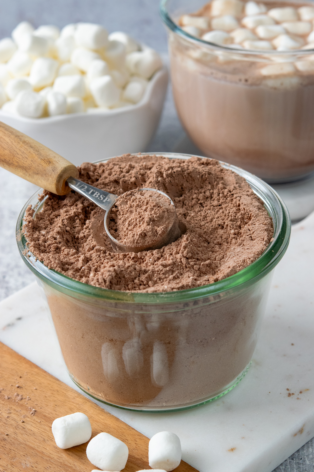 Homemade hot chocolate mix in a jar with a scoop, alongside a mug of hot chocolate and a bowl of mini marshmallows.