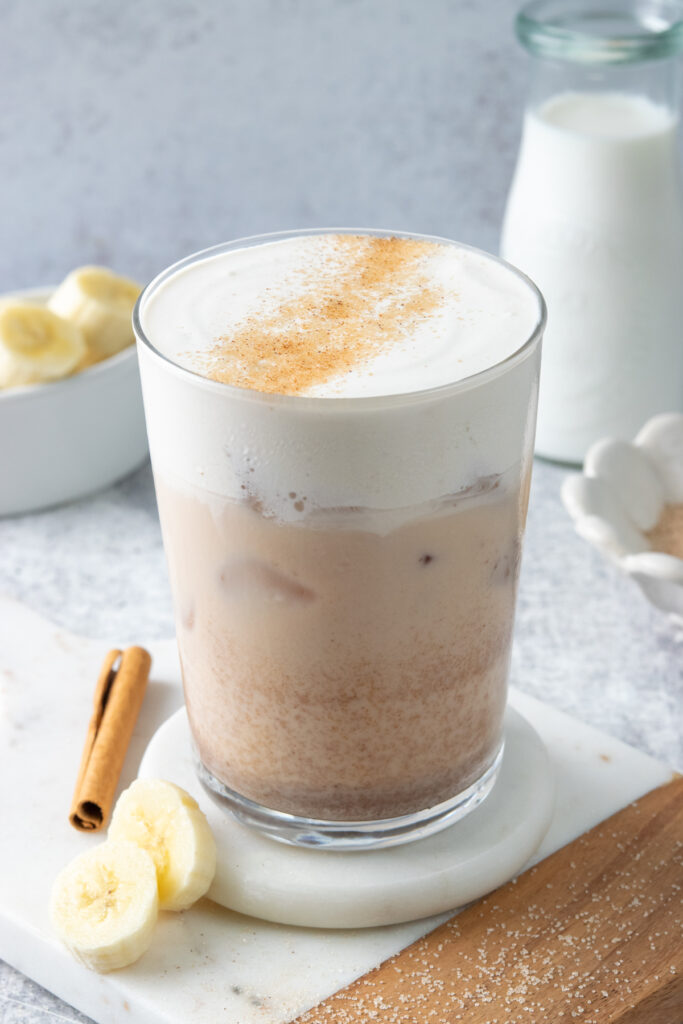 Iced banana bread chai latte topped with creamy banana bread cold foam and cinnamon sugar, with sliced bananas and cinnamon sticks alongside the Starbucks copycat drink.