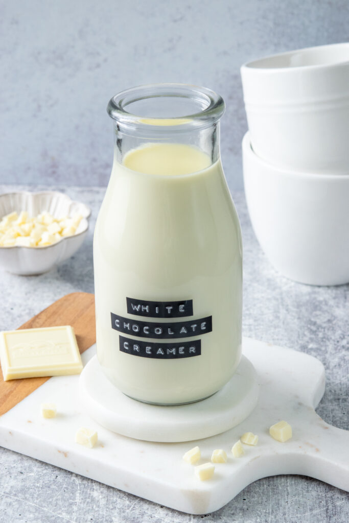 Bottle of homemade white chocolate coffee creamer surrounded by chopped white chocolate, a white chocolate bar, and white coffee mugs.