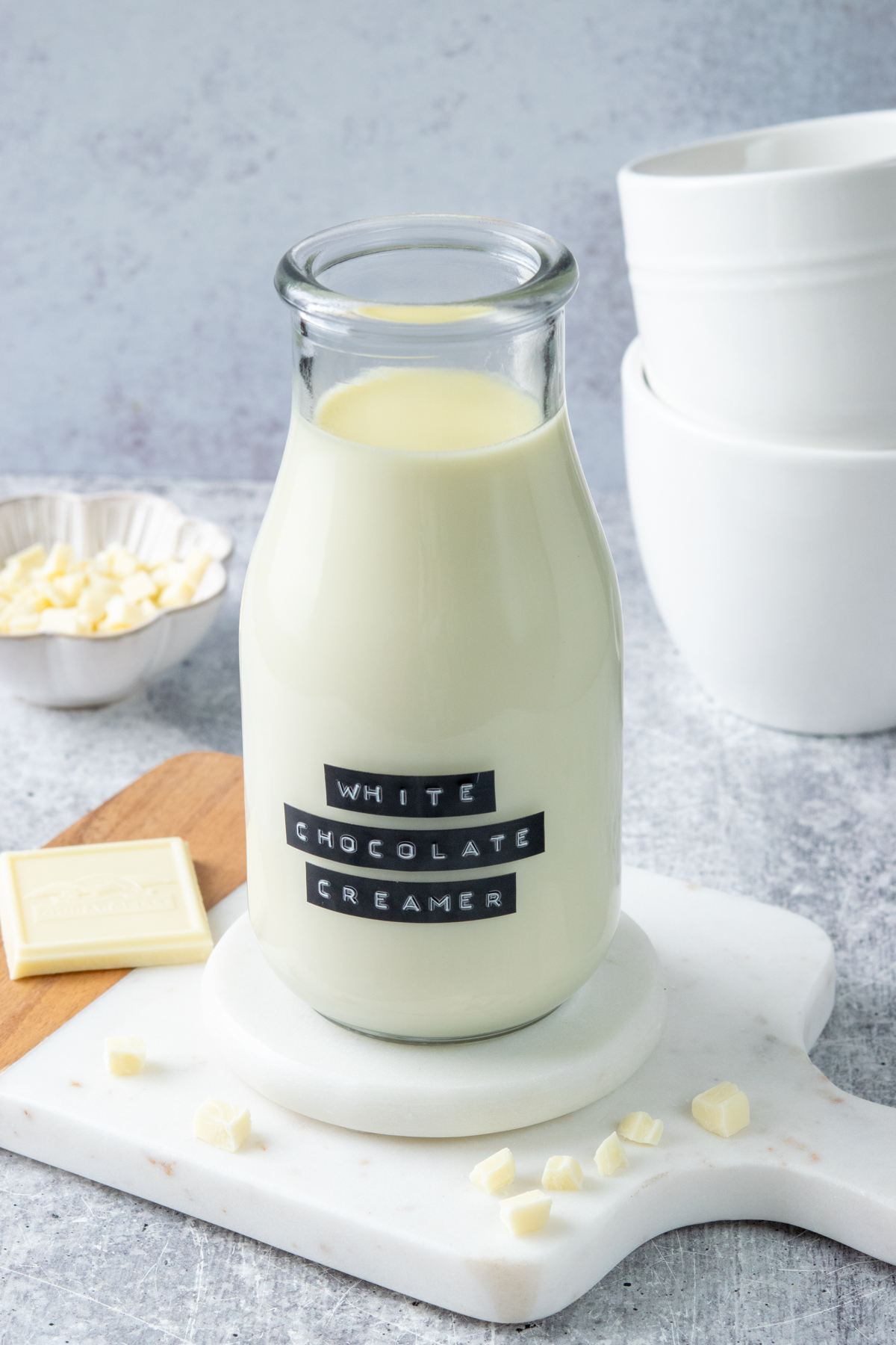 Bottle of homemade white chocolate coffee creamer surrounded by chopped white chocolate, a white chocolate bar, and white coffee mugs.
