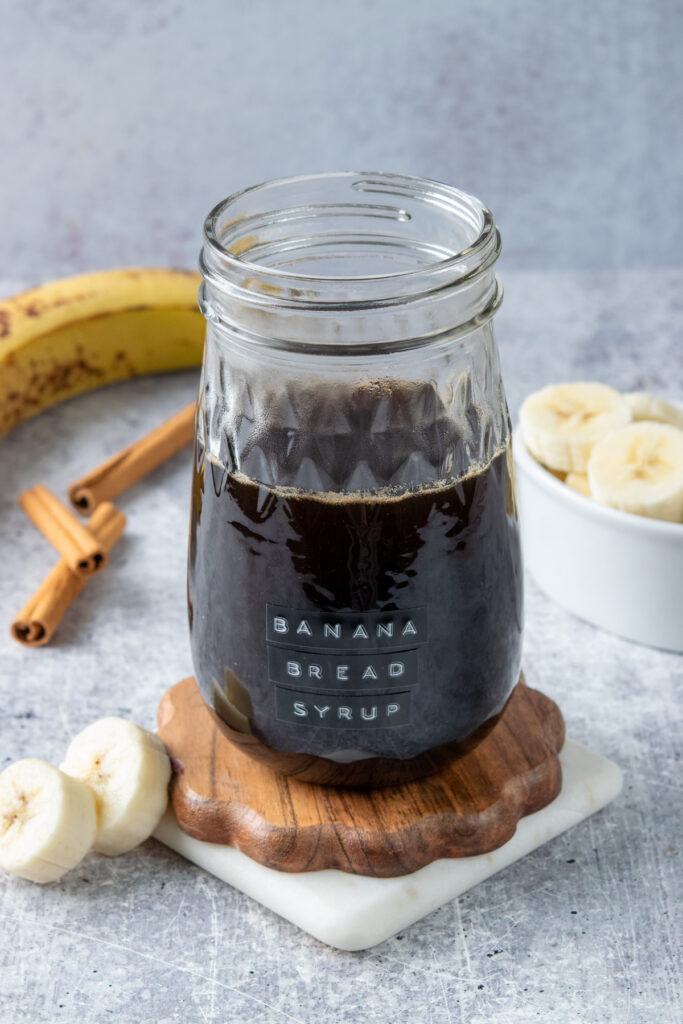 Jar of homemade banana bread syrup labeled “banana bread syrup,” surrounded by sliced bananas, a ripe banana, and cinnamon sticks.