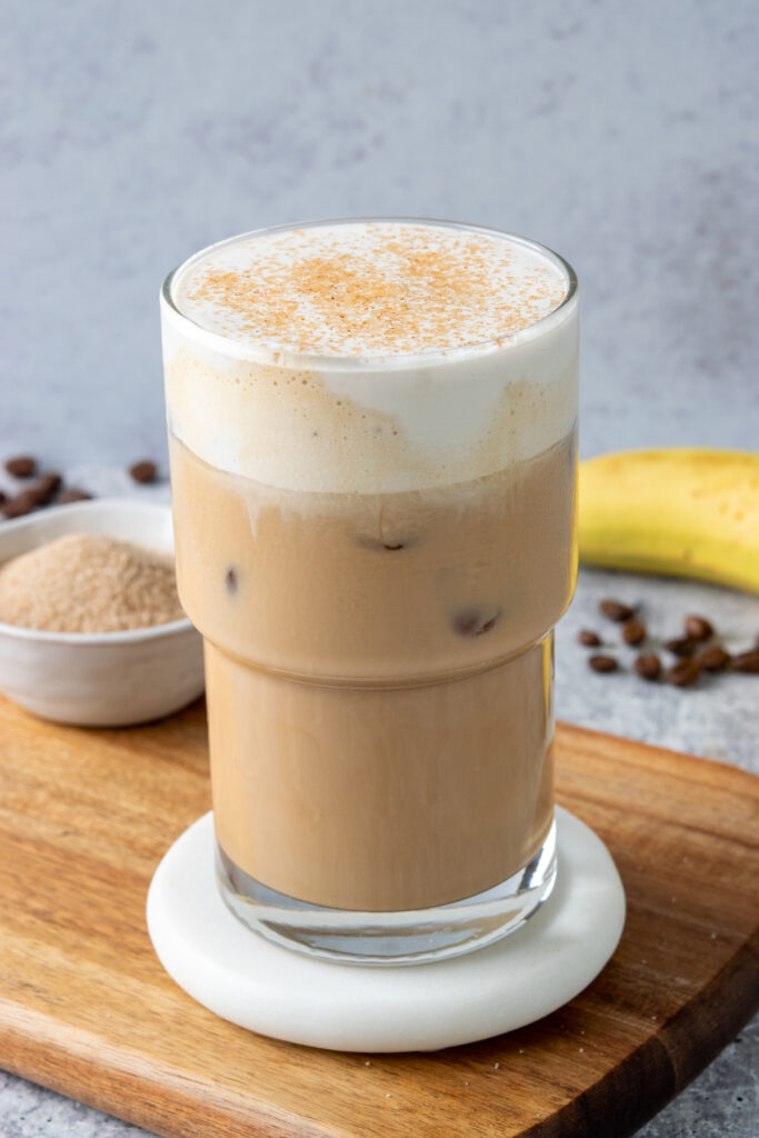 Starbucks copycat iced banana bread latte topped with creamy banana bread cold foam and cinnamon sugar, served with a banana, espresso beans, and a small bowl of cinnamon sugar.