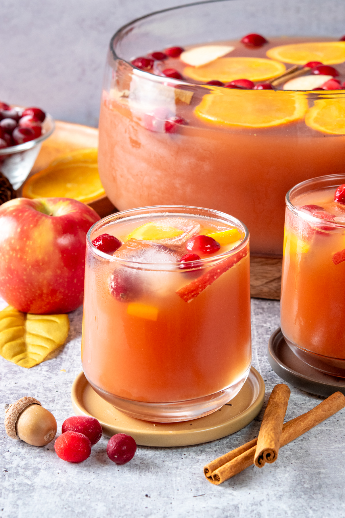 Thanksgiving punch made with apple cider, cranberry juice cocktail, orange juice, and ginger ale, served in a large punch bowl and glasses with cranberry, orange, apple, and cinnamon stick garnishes.